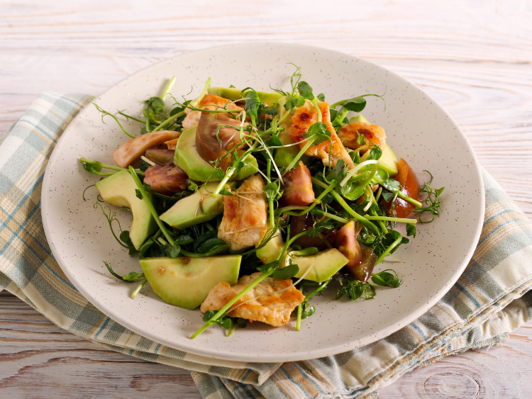A fresh salad of grilled chicken slices, avocado, tomato wedges, and microgreens on a speckled ceramic plate over a striped cloth napkin.
