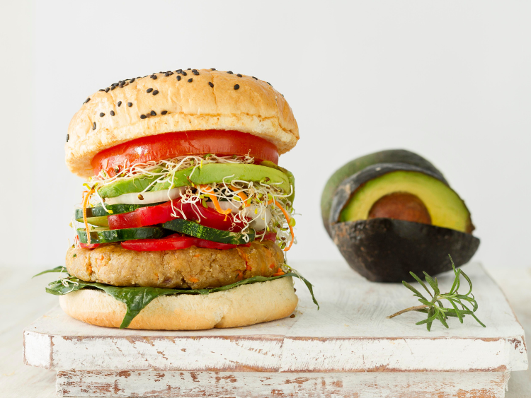 Healthy veggie burger with avocado slices, sprouts, tomato, cucumber, bell pepper, and a plant-based patty on a sesame seed bun, served on a rustic wooden board with a halved avocado.