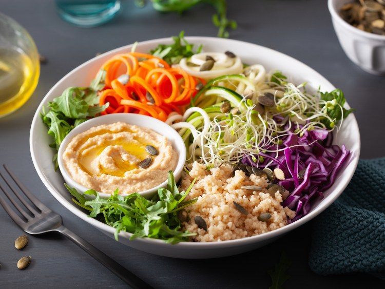 Vegan couscous lunch bowl with spiralized carrots and zucchini, shredded purple cabbage, fresh arugula, crunchy sprouts, creamy hummus dip, and pumpkin seeds, drizzled with olive oil for a healthy plant-based meal.